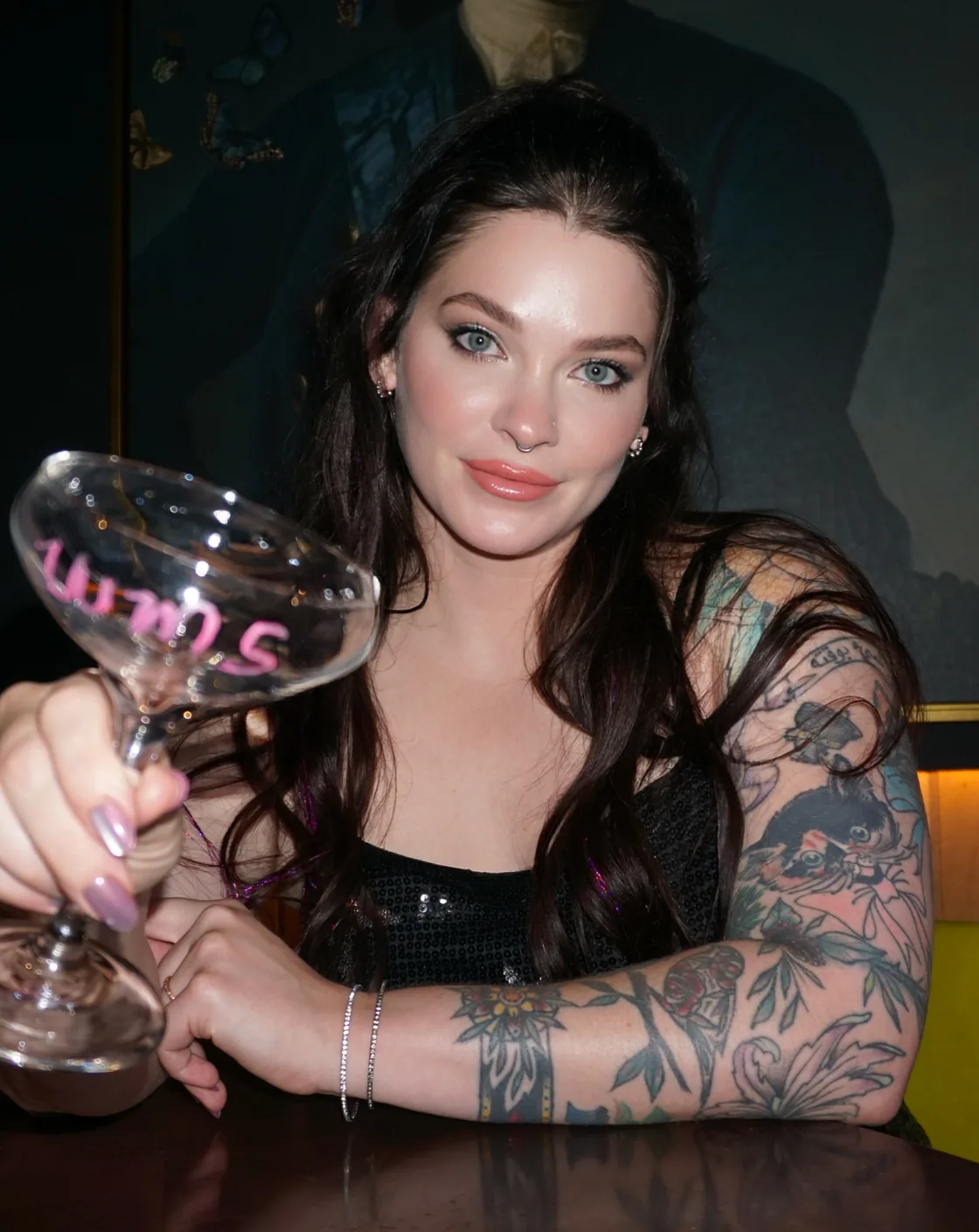 A guest with long dark hair and a tattooed arm sitting in a dark restaurant booth, raising a coupe glass toward the camera